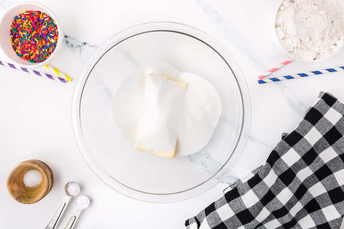 sugar and butter in a clear glass mixing bowl with sprinkles nearby